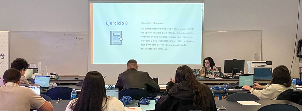 Foto en el aula durante el taller. De espaldas 6 estudiantes sentados trabajando en computadoras. De frente María dando la clase acompañada de una presentación proyectada con el título &ldquo;Ejercicio 8&rdquo;
